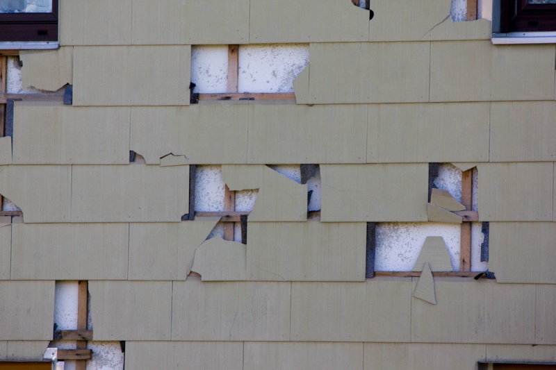 A close up of yellow siding on a home with several cracks in it and tiles missing. The white interior of the house can be seen through the cracks.